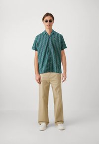 Cămașă cu mâneci scurte, cu modele verzi și albastre, cu forme geometrice, asortată cu pantaloni bej și adidași albi. Ochelari de soare rotunzi, închiși la culoare, purtați.