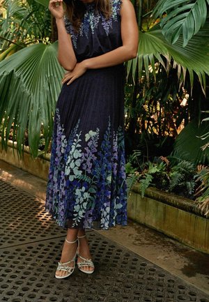 Bloemenjurk met een donkere achtergrond met paarse en groene plantenpatronen; lichte stof, vloeiende silhouet en knielange snit. Bruine sandaaltjes met banden.