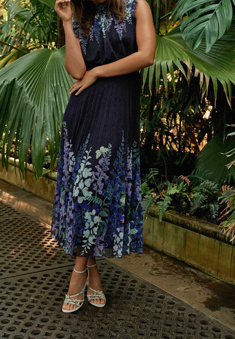 Robe florale avec un fond sombre comportant des motifs de plantes violettes et vertes ; tissu léger, silhouette fluide et coupe à mi-genoux. Sandales à talons bruns.