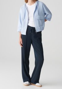 Žena v světle modré krátké mikině na zip, bílém tričku, tmavě modrých širokých kalhotách se stahovací šňůrkou a bílých teniskách s béžovou podrážkou.