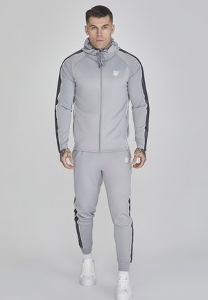 Grijze trainingsset met zwarte zijstripe, een ritsjack en elastische broek. Bevat een capuchon en witte logo-detail op de borst. Witte sneakers.