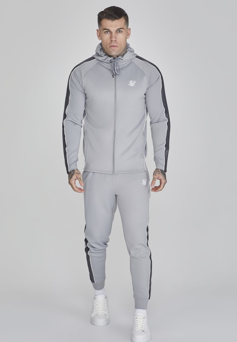Grå träningsoverall med svarta sidostråkar, zip-up jacka och byxor med elastisk midja. Har en huva och vitt logodetalj på bröstet. Vita sneakers.