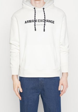 Hvid hættetrøje med sort "ARMANI EXCHANGE" tekst og stribede snore, båret med blå jeans, viser overkrop og arme.