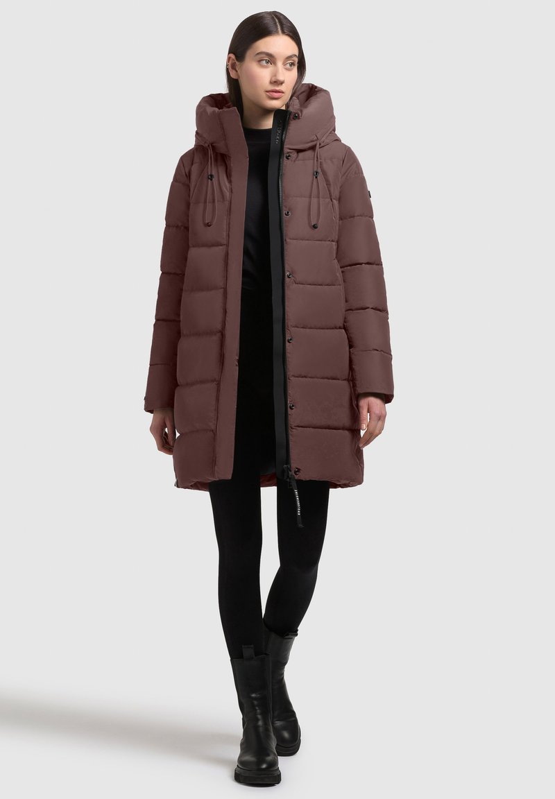 Brun pufferskåp med huva, med en quiltad design, framknappar, svarta accenter och dragsko-detaljer. Bärs med svarta leggings och stövlar.