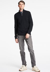 Schwarzer, mit Reißverschluss versehenen Pullover mit weicher Textur, kombiniert mit hellgrauen, ihn abgewetzten Jeans und cremefarbenen Sportschuhen. Das Model steht aufrecht vor einem weißen Hintergrund.