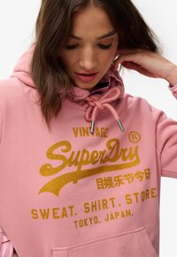 Roze hoodie gemaakt van zacht materiaal, met gouden rhinestone tekst: "Vintage SuperDry," voorzien van een capuchon met trekkoord en een kangoeroezak.