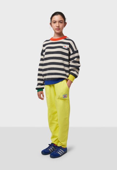 Suéter de punto a rayas en azul marino y blanco con acentos en rojo y amarillo, combinado con pantalones de chándal amarillos brillantes y zapatillas deportivas azules.