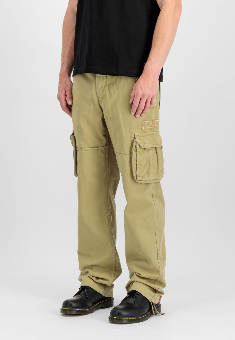 Mann trägt khakifarbene Cargohosen mit großen Seitentaschen und schwarze Schnürschuhe, steht vor einem schlichten hellen Hintergrund.