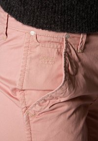 Ljusrosa byxor med en slät textur, prydda med subtila sömdetaljer och en liten ficka som accentueras av ljusbeige tråd.