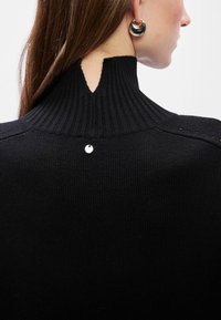 Μαύρος πλεκτός σγουρός με ψηλό ριμπ λαιμό, που διαθέτει μια V-σχήματος κοπή στο πίσω μέρος και μια μικρή ασημένια λεπτομέρεια.