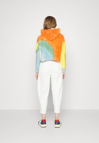 Felpa con cappuccio corta in motivo tie-dye con tonalità di arancione, blu e giallo, abbinata a pantaloni bianchi ampi e sneakers vivaci.