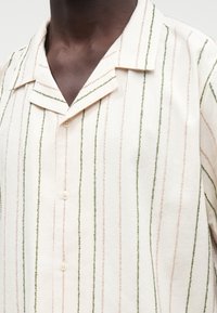 Tamsios odos asmuo, vilkintis kreminę marškinių su sagomis, su vertikaliomis žaliomis ir smėlio spalvos tekstūruotomis juostelėmis bei kempingo tipo apykakle.