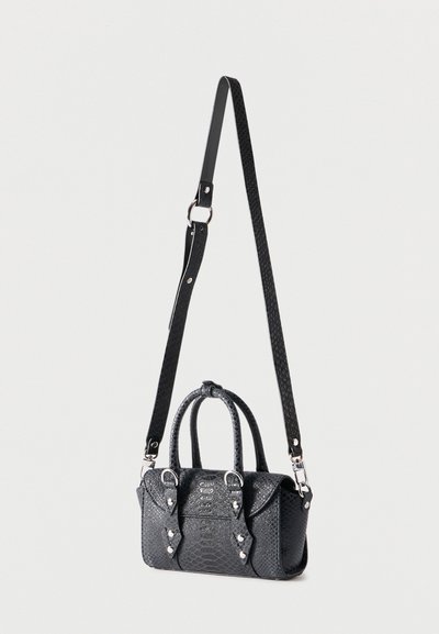 Μαύρη τσάντα χειρός από δερματίνη με υφή, με κοντές λαβές και μακριά ρυθμιζόμενη λουρίδα ώμου, κρεμασμένη σε απλό φωτεινό φόντο.