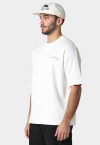 Camiseta de algodón blanca con mangas cortas, que presenta un sutil texto bordado "PAS DE MONACO" en dorado. Complementada con una gorra beige claro.
