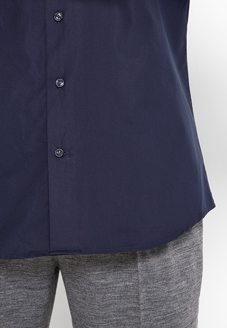 Granatowa koszula zapinana na guziki o gładkiej fakturze i zaokrąglonym dole, zestawiona z szarymi spodniami o fakturze, z widocznymi guzikami i detalami szycia.