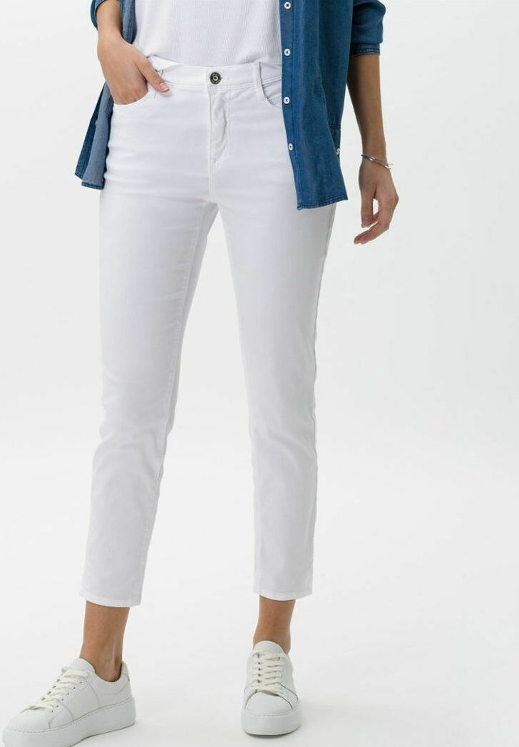 Personne portant un jean blanc slim, des baskets blanches, un haut blanc et une chemise en denim bleu, une main dans la poche.