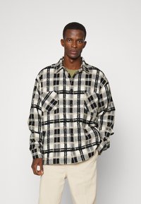Camisa xadrez em preto e branco feita de tecido macio, com mangas longas, dois bolsos no peito e fechos de botão, combinada com calças creme.