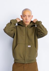 Sweatshirt com fecho éclair na cor verde oliva, com um bolso frontal, bainha e punhos canelados, apresentando um pequeno patch de logótipo perto do fecho. Material de fleece suave.