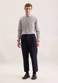 Geruit overhemd met knopen in wit en zwart, gecombineerd met marineblauwe broek met hoge taille. Model draagt zwarte leren schoenen en staat in een neutrale omgeving.