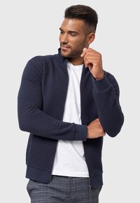 Mørkeblå lynlåsjacket i tekstureret stof, med ribbede manchetter og krave; båret over en hvid T-shirt, parret med mønstrede bukser.