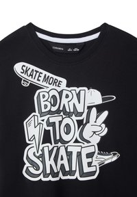 T-shirt in cotone nero con testo bianco in grassetto: "NATO PER SKATEARE" insieme a grafiche di skateboard e un simbolo di pace, scollo tondo e maniche corte.