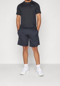 Camiseta atlética negra de manga corta, combinada con pantalones cortos oscuros. Ambos están hechos de un material que absorbe la humedad. Se lleva con zapatillas blancas.