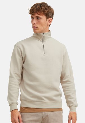 Mann trägt beige Viertel-Reißverschluss-Pullover und braunes Unterhemd, schaut nach links, steht vor einfarbigem Hintergrund.