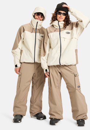 Beige en crème ski-jassen met capuchons, ritsen aan de voorkant en zakken; gecombineerd met beige broeken; beide hebben zwarte accenten en stevige materialen.