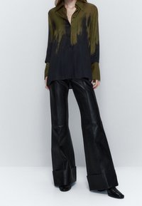 Femme portant une chemise boutonnée oversize olive et noire, associée à un pantalon en cuir noir à jambes larges et des chaussures noires, sur fond gris uni.