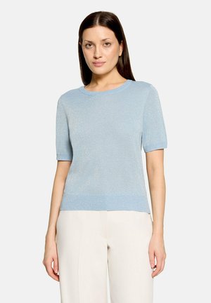 Femme aux cheveux bruns lisses portant un haut bleu clair à manches courtes et un pantalon crème, debout devant un fond uni et clair.