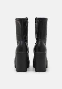 Botas de tobillo de cuero negro con punta redonda, tacón cuadrado alto, cremalleras laterales y un acabado liso. La vista trasera muestra los detalles de las costuras y la silueta.