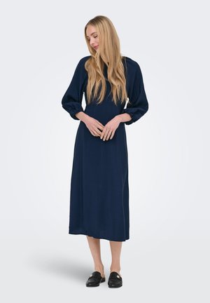 Femme aux longs cheveux blonds portant une robe midi bleu marine avec des manches bouffantes et des mocassins noirs sans lacets, debout devant un fond blanc.