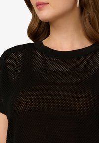 Černý pletený top s síťovaným designem, s kulatým výstřihem a krátkými rukávy. Textura je otevřená a prodyšná, ideální pro vrstvení.