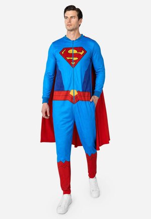 Homme debout portant une combinaison bleue et rouge sur le thème de Superman, avec une cape rouge et des baskets blanches sur un fond uni.