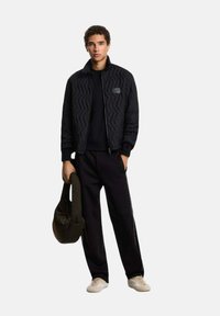 Jonge man in een zwarte gewatteerde jas, zwarte broek en witte sneakers, met in één hand een zwarte gewatteerde tas, staand tegen een witte achtergrond.