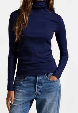 Frau trägt einen marineblauen, gerippten Rollkragenpullover und hellblaue Jeans, eine Hand in der Tasche, vor einfachem Hintergrund.