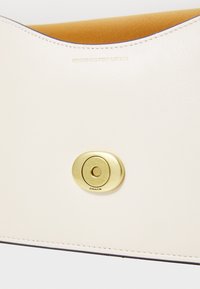 Coated leather handbag in light cream with a smooth texture, featuring a gold circular closure and embossed branding.