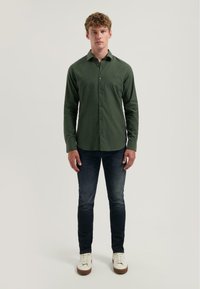 Donkergroen overhemd met lange mouwen en subtiele textuur, gecombineerd met donkerblauwe slim-fit jeans en witte sneakers. Casual outfit presentatie.