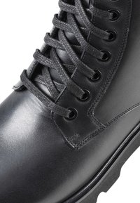 Bota de cuero negro con acabado texturizado, que presenta cordones negros redondos, ojales de metal y un diseño de suela resistente para mayor tracción.