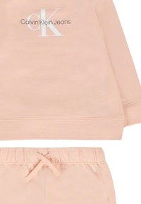 Jasnoróżowy dres składający się z bluzy z długim rękawem i spodni z elastycznym pasem. Bluza ma biały logo, natomiast spodnie mają sznurek do regulacji.
