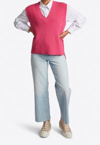 Rosa gestrickte Weste mit V-Ausschnitt, kombiniert mit hellblauen weit geschnittenen Jeans und beigen Sneakers. Strukturierter Stoff mit seitlichen Schlitzen für einen stilvollen Look.