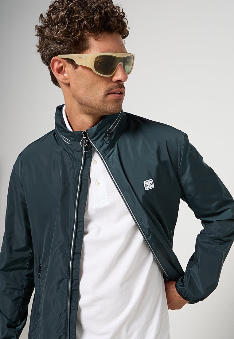 Giacca leggera verde scuro con zip frontale, dotata di colletto alla coreana e un piccolo logo. Indossata sopra una polo bianca.