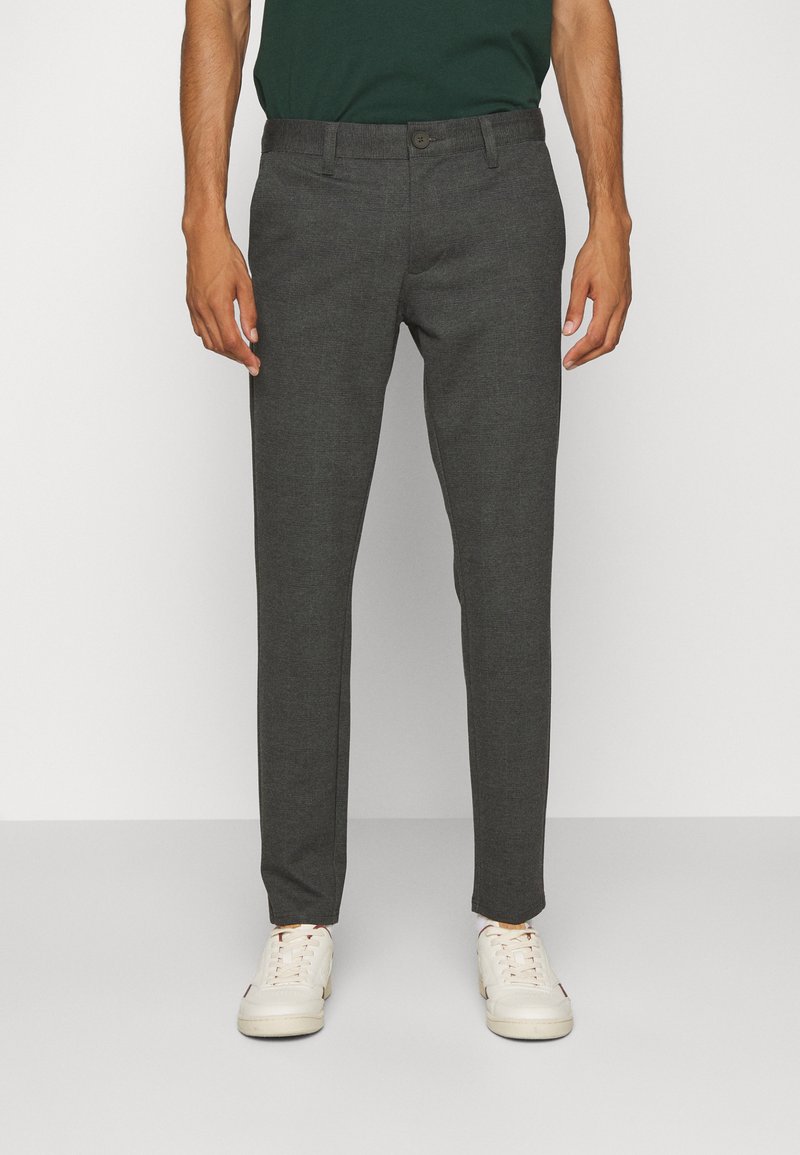 Pantalones oscuros ajustados en gris con un diseño entallado, cierre de botón y trabillas para cinturón. Elaborados en una tela texturizada, combinados con zapatillas blancas.