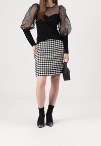 Svart långärmad topp med genomskinliga, puffärmar prydda med pärlor; kjol i hundtandsmönster; svart handväska; svarta ankelsockor med klackar.