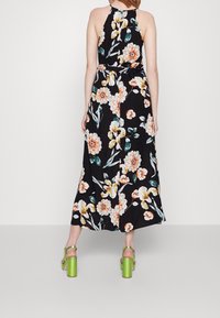 Robe maxi fleurie avec un fond noir orné de fleurs orange et blanches, col halter et jupe fluide ; associée à des talons carrés verts.