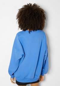 Osoba s kudrnatými vlasy, která má na sobě oversized modrou mikinu a černou sukně, stojí otočená zády k prostému bílému zdi.