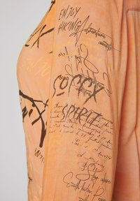 Oranges Stoffhemd mit verschiedenen handschriftlichen Stilen und Mustern in Dunkelbraun, darunter Phrasen und abstrakte Designs.