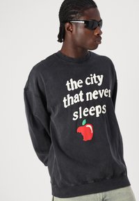 Black, oversized sweatshirt with white text reading "the city that never sleeps" and a red apple graphic with a bite taken out.