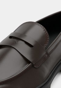 Brune lærloafers med en glat overflade, der har et udskæringsdesign og subtil syning langs kanten, monteret på en sort sål.