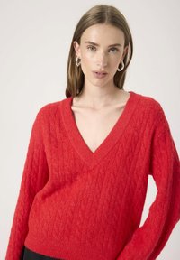Woman with light skin and straight brown hair wears silver hoop earrings and a bright red V-neck cable knit sweater, posing against plain background.